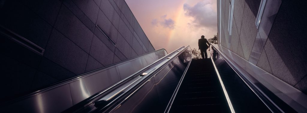 A man at the top of an escalator with a rainbow in the sky above him.