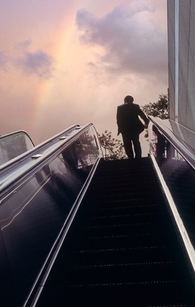 A man at the top of an escalator with a rainbow in the sky above him.