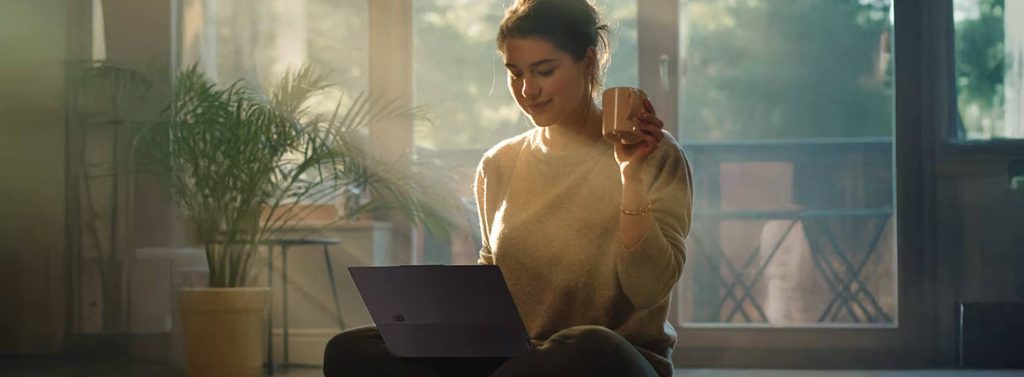 A woman sitting on her floor at home, using her laptop and holding a cup of coffee as the sun shines through her windows.